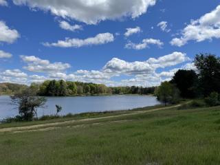 View of Trail Lake from paved asphalt trail. Blue sky, white clouds, trees, and kayak launch in view.
