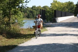 A bike rider faces rides toward us on a limestone trail flanked by green foliage. Water is seen behind the rider. 