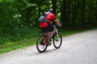 Cyclist with red shirt and gray backpack on limestone trail with green trees in background