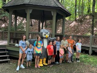 A group of smiling kids pose with an owl mascot near a wooden gazenbo. 