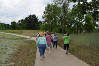 A group of hikers walks away from the front of the photo along a paved, curved trail along green meadow and trees