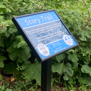 A framed sign contains a blue poster designating the story trail start in front of green foliage 