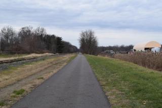 View down dark gray asphalt paved trail  with train tracks on the left and grass on the right