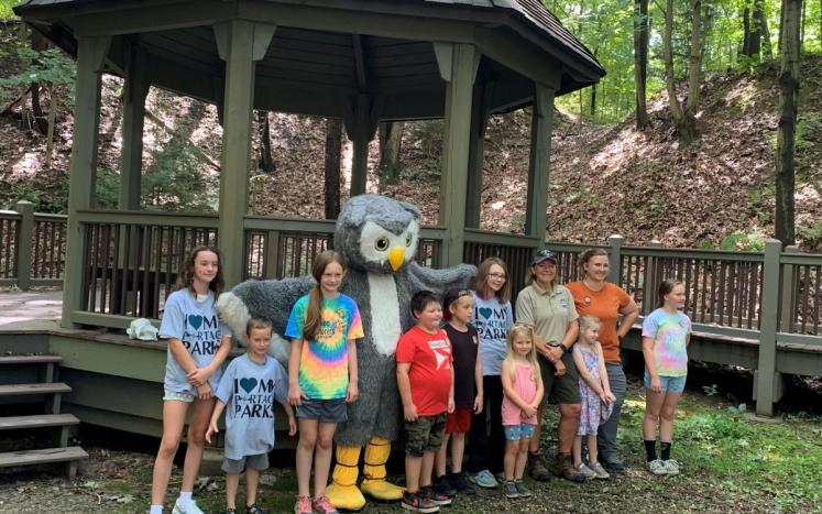 A group of smiling kids pose with an owl mascot near a wooden gazenbo.