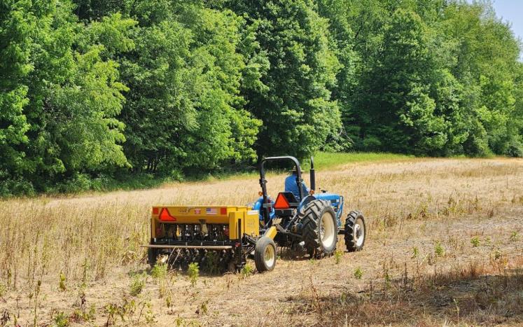 blue tractor pulling yellow seed drill across a field with green trees in background